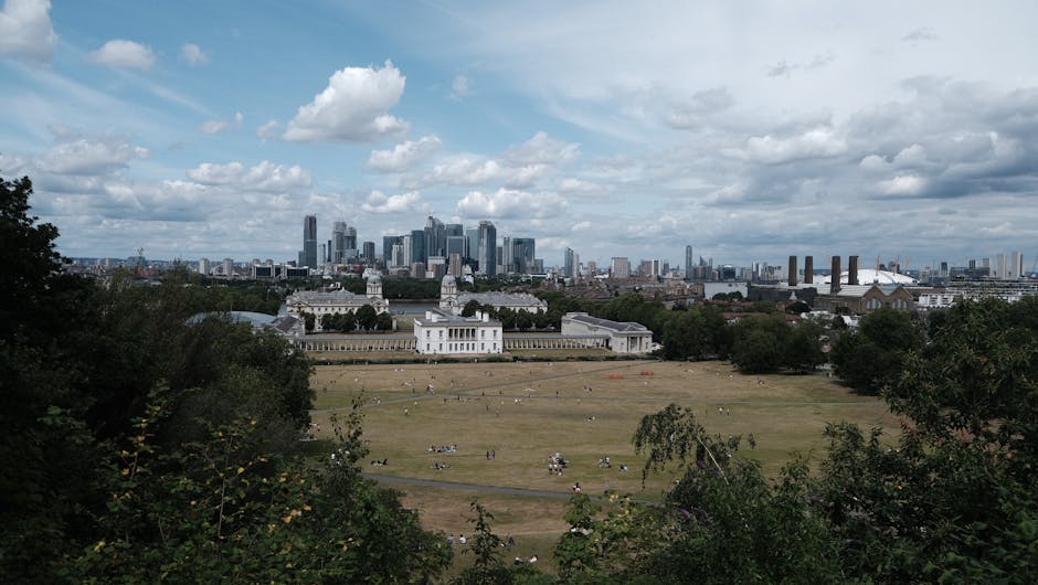 A panoramic view of Greenwich Park with a well-maintained lawn in the foreground, where small groups of people are sitting and relaxing. The park features a neatly trimmed grassy area with areas of ex