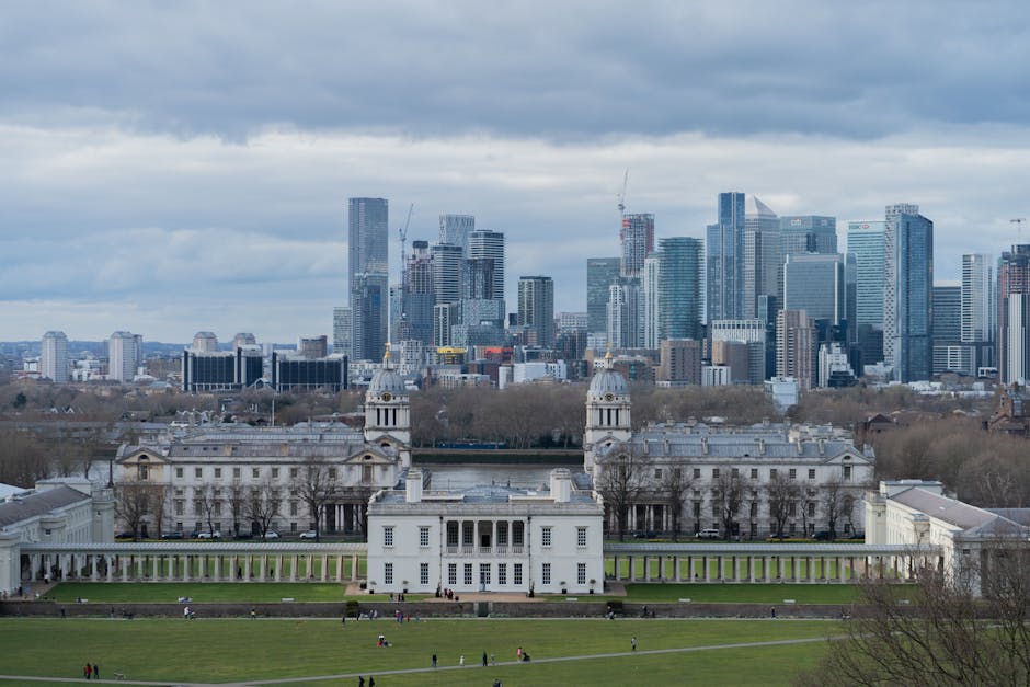 A panoramic view of Greenwich Park with a well-maintained lawn in the foreground, where small groups of people are sitting and relaxing. The park features a neatly trimmed grassy area with areas of ex